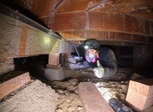 A worker in a crawlspace, wearing protective gear, inspects a concrete footing and post supporting a beam